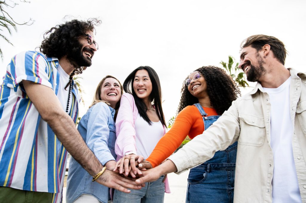 Diverse group of cheerful millennial friends standing together stacking hands in the street