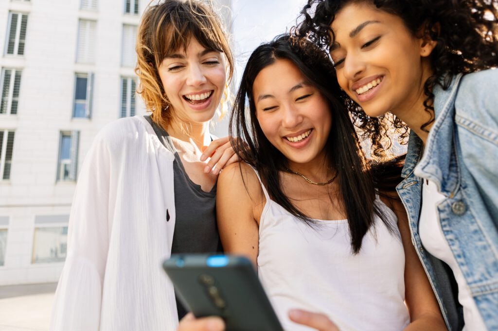 Young group of diverse girl friends watching at mobile phone screen outdoors.